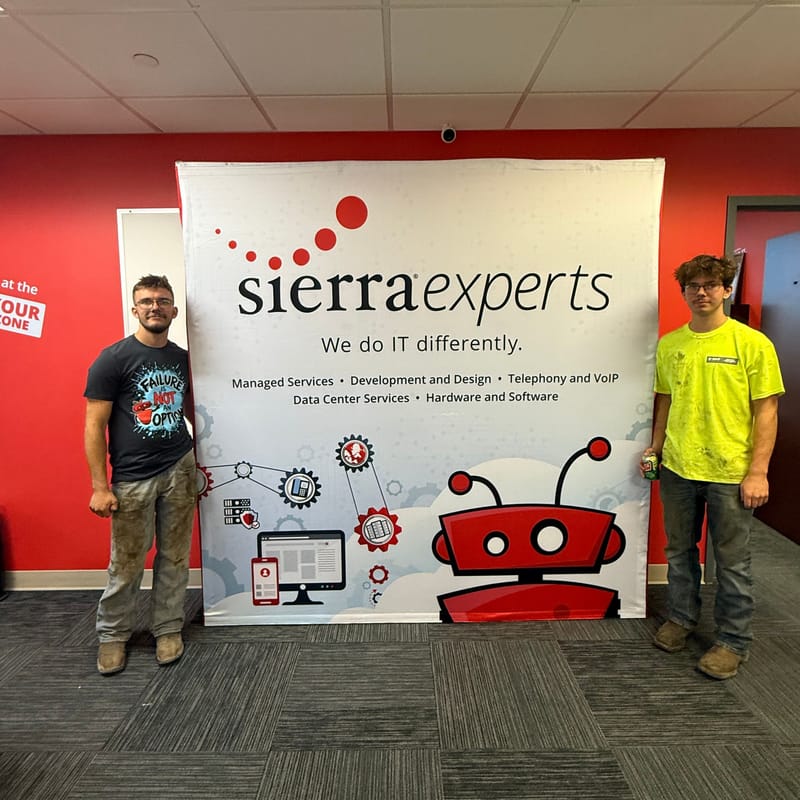 Two team members beside the Sierra Experts “We do IT differently” banner with the red robot mascot